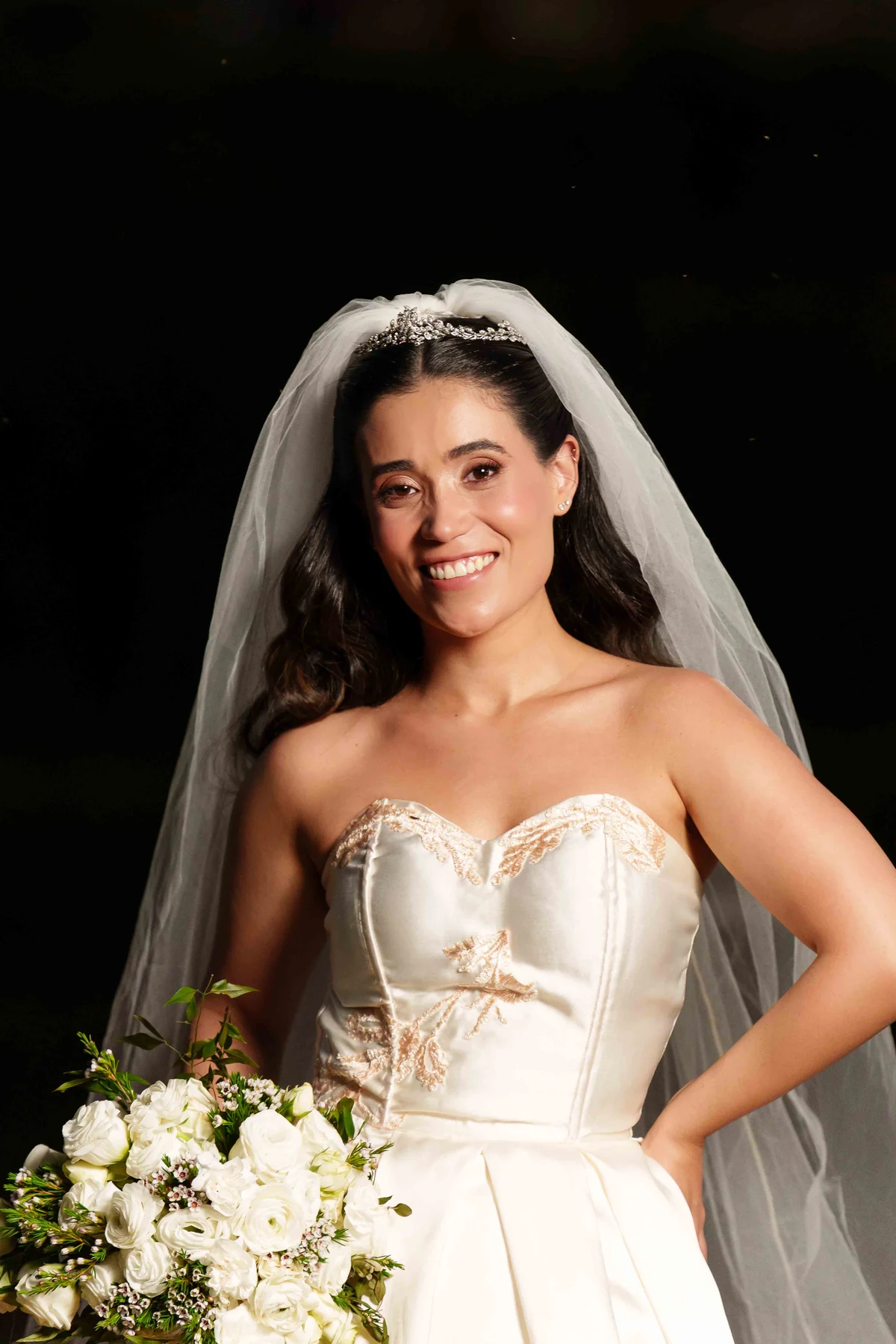 Gabi no dia do casamento, sorrindo com seu vestido, véu e buquê de flores brancas
